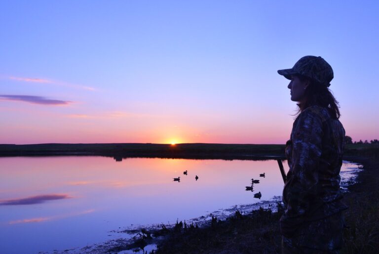 Waterfowl hunter silhouetted at sunrise beside decoys on a marsh pond